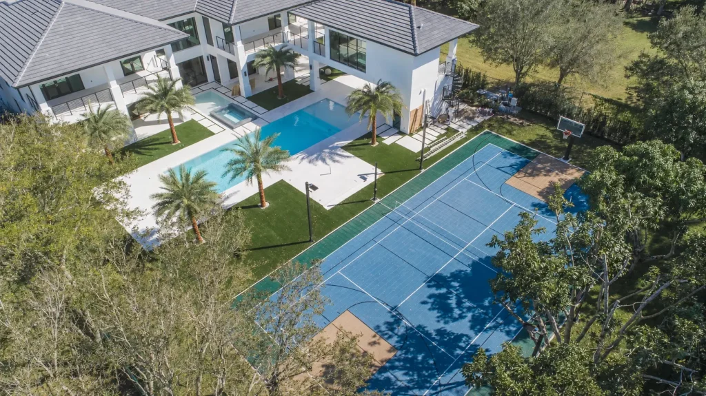 Aerial view of a blue and green residential tennis court beside a luxury home and swimming pool